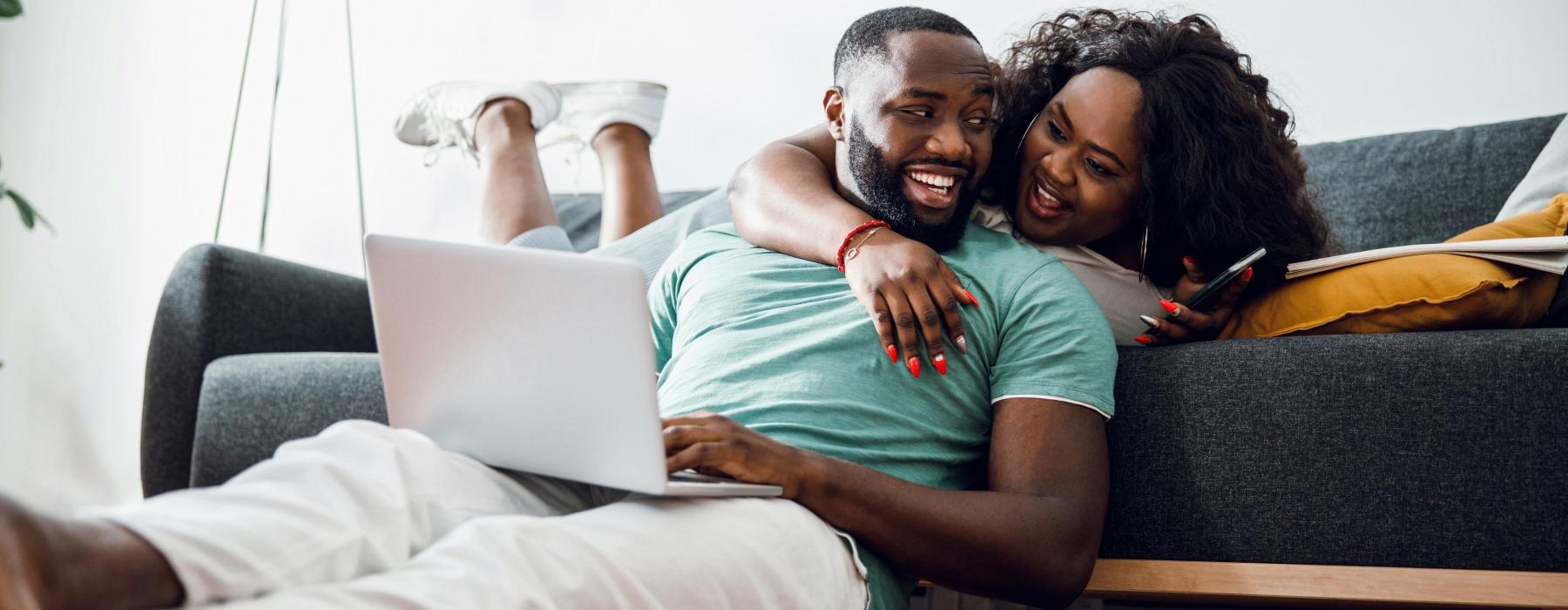 a man and woman laughing while sitting on a couch with a laptop