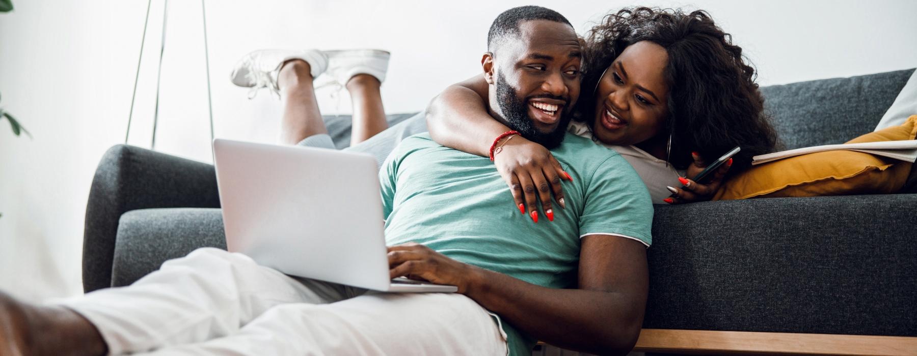 a man and woman laughing while sitting on a couch with a laptop